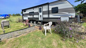 A recreational vehicle parked in a grassy area with a small patio setup including a white chair and a table.