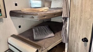 Een compact RV-interieur met een slaapstapelbed, een zithoekje en een raam met zonwering.