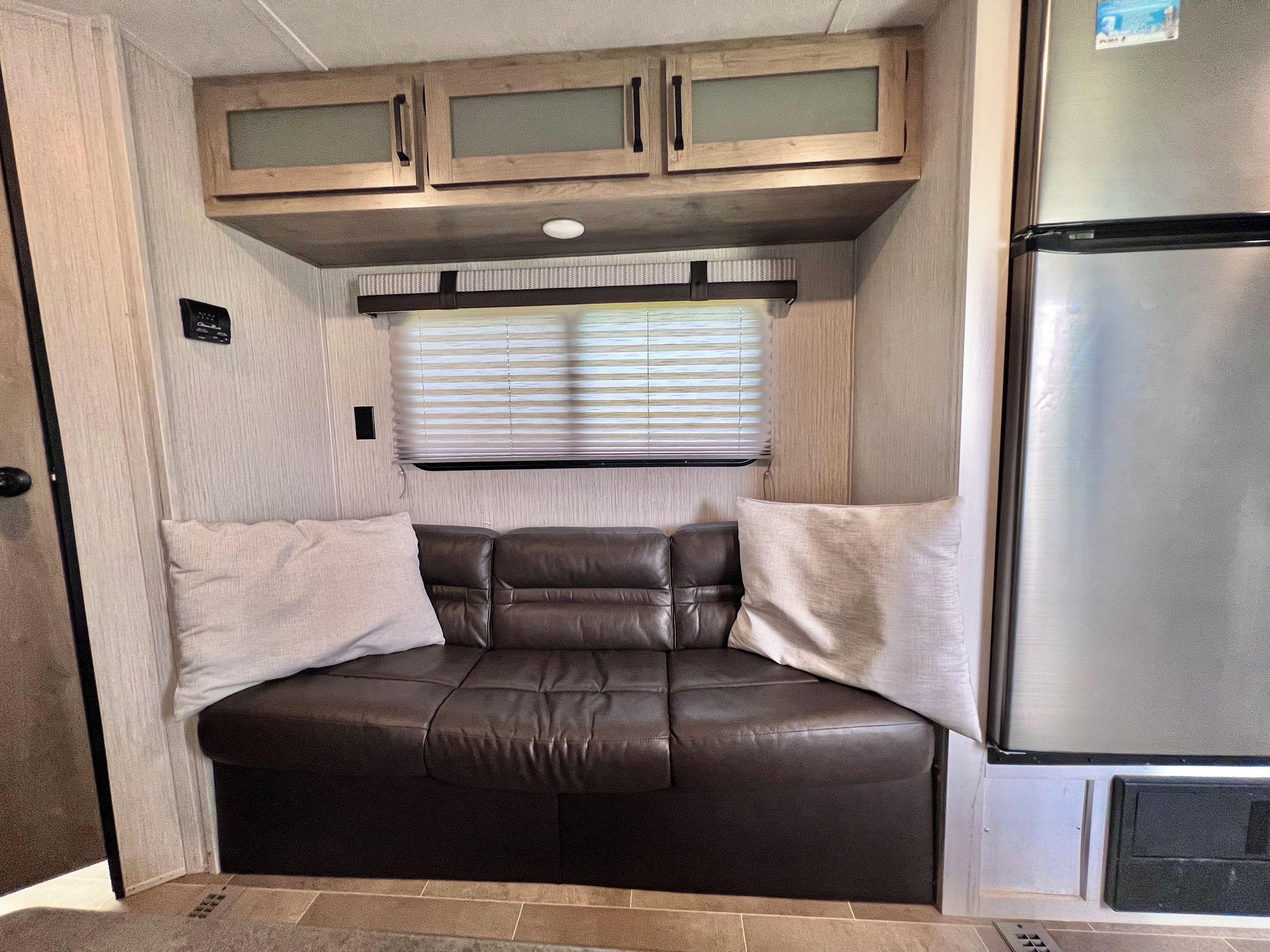 A compact RV interior with a kitchenette, a cushioned seating area, and a window with blinds.