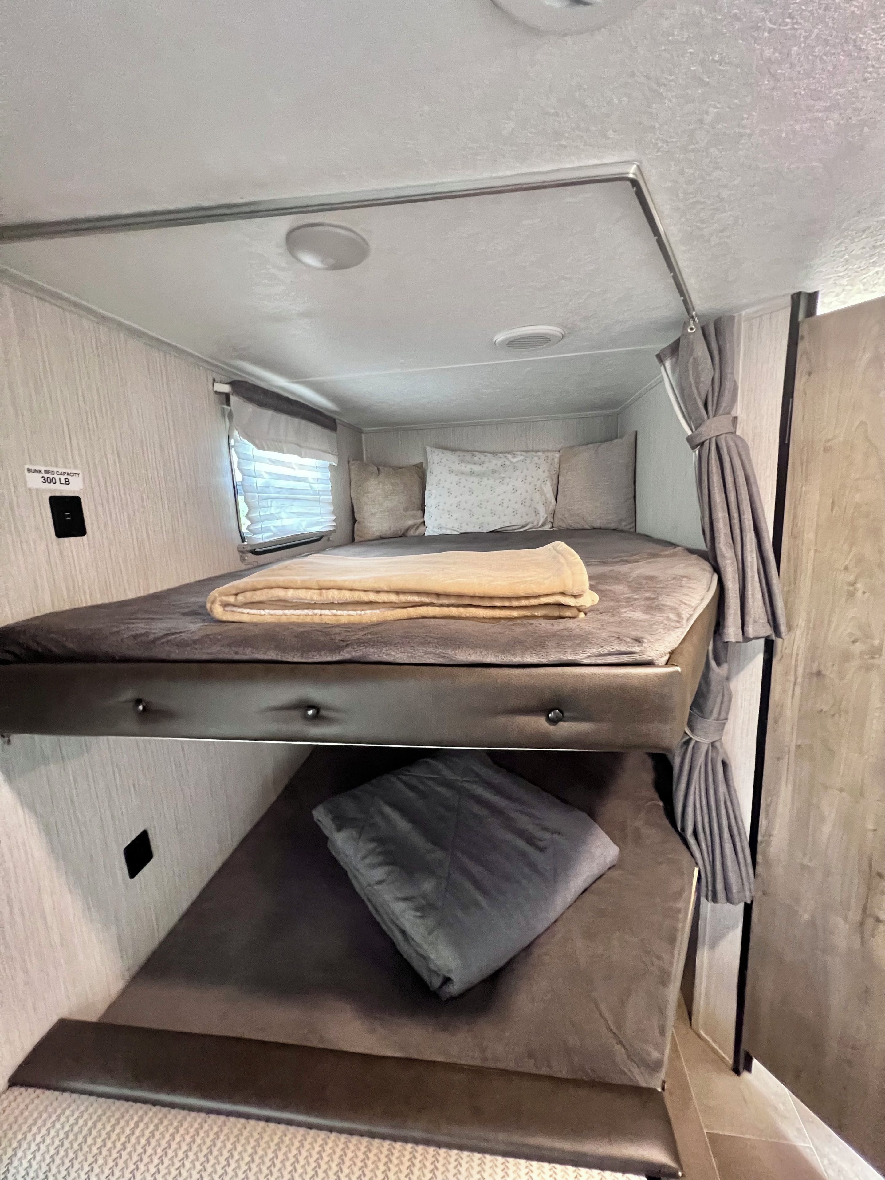 Un lit superposé avec un matelas et des oreillers, une fenêtre avec des stores et un panneau indiquant une capacité de charge maximale de 300 livres pour le lit superposé.