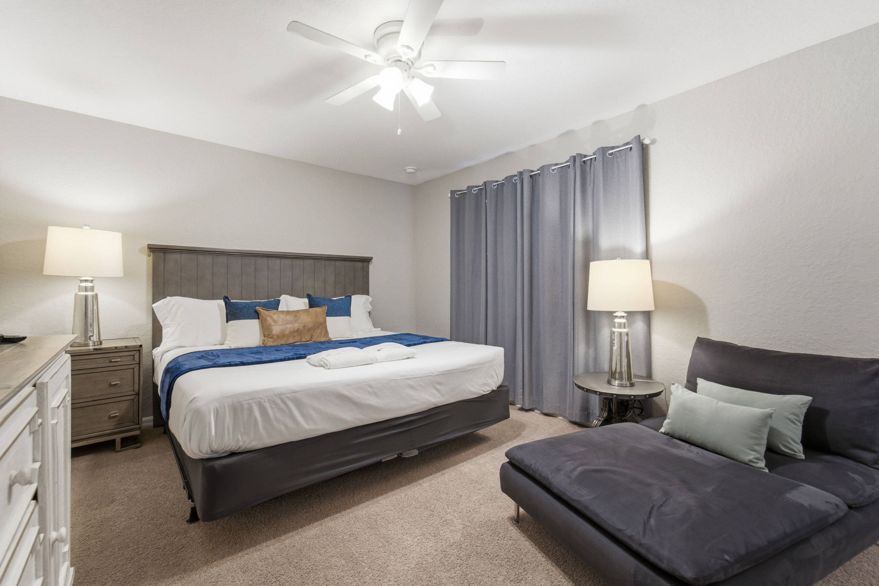 A hotel room with a bed, a desk, a chair, a ceiling fan, and curtains.