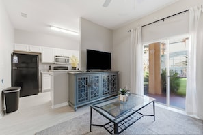Een moderne keuken met witte kastjes, een zwarte koelkast, een magnetron en een fornuis. Er staat een glazen tafel met een plantje op, en een schuifpui die leidt naar een balkon met uitzicht op groen.