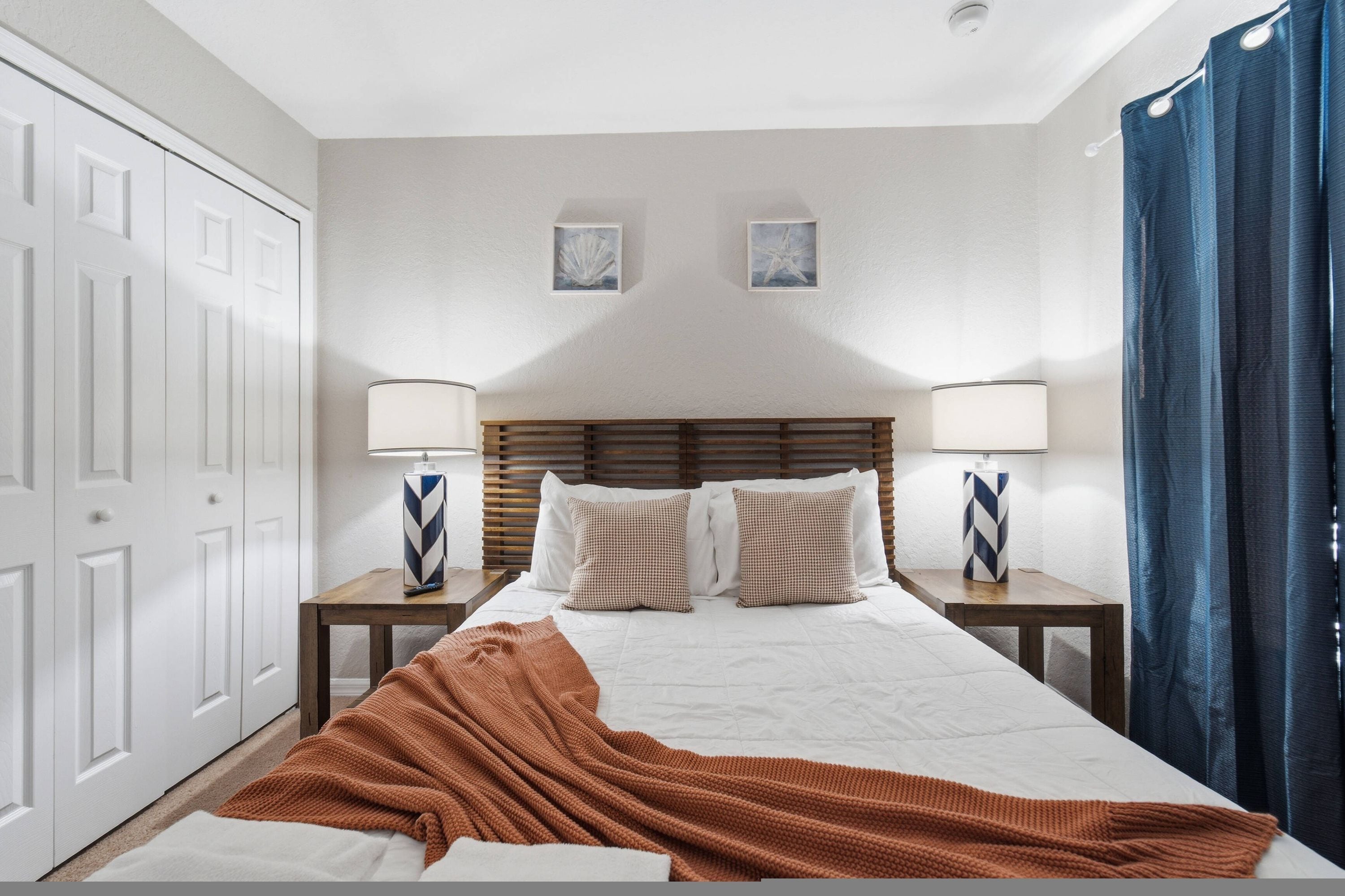 A hotel room with a bed, two bedside tables, a wooden headboard, a brown throw blanket, and a window with blue curtains.