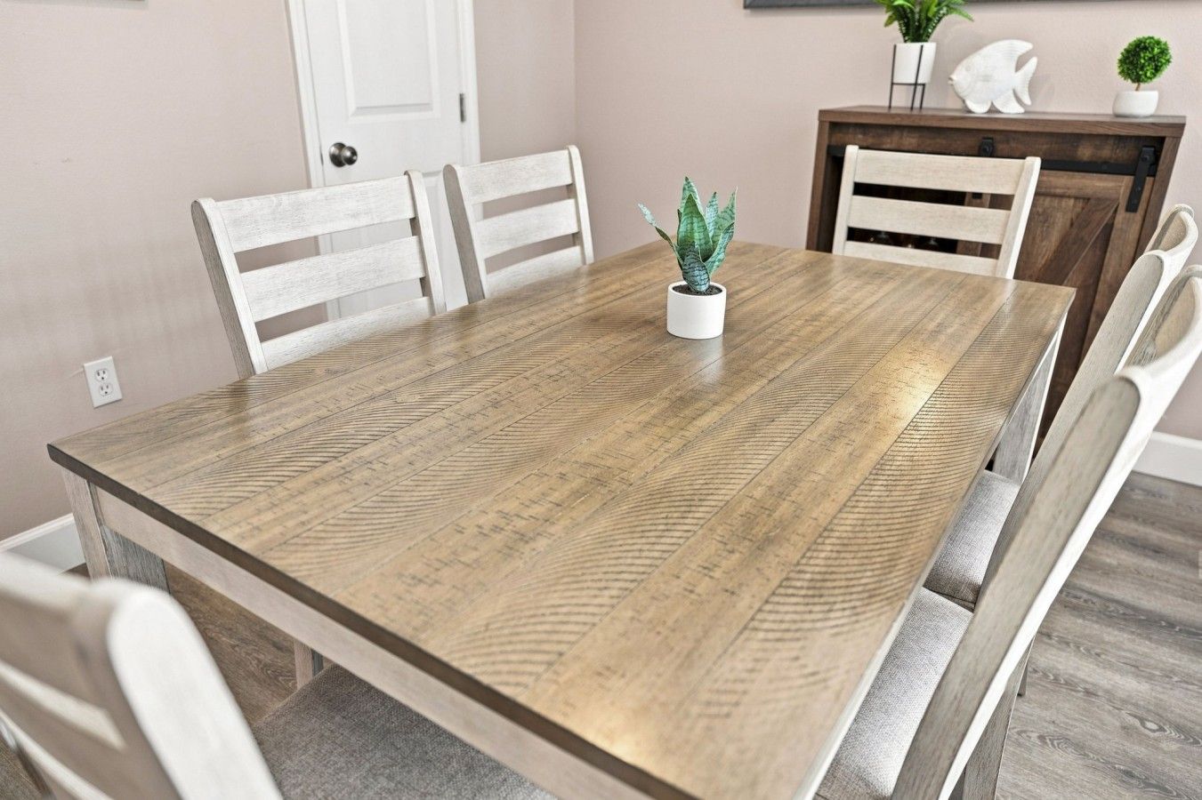 A dining table with a light wood finish, surrounded by white chairs, in a room with a white door and a small potted plant on the table.