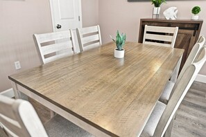A dining table with a light wood finish, surrounded by white chairs, in a room with a white door and a small potted plant on the table. - Modern 2BR Condo in a Prime Walkable Location (Madeira Beach)