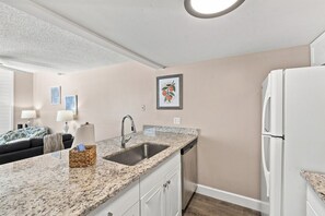 A compact kitchen with a granite countertop, stainless steel sink, and a white refrigerator. - Modern 2BR Condo in a Prime Walkable Location (Madeira Beach)