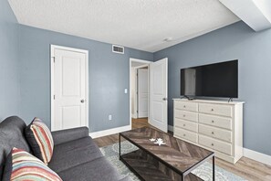 A living room with a sofa, a coffee table, a TV on a dresser, and a door leading to another room. - Modern 2BR Condo in a Prime Walkable Location (Madeira Beach)