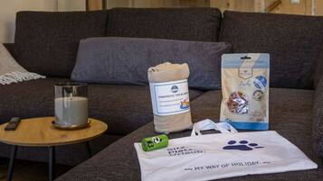 A living room with a grey sofa, a glass candle holder, a rolled-up blanket, a pet food bag, and a tote bag with a paw print.