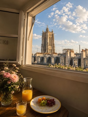 Vista per la finestra d'una torre històrica, una taula amb un plat de fruita, un ram de flors i un got de suc d'aranja.