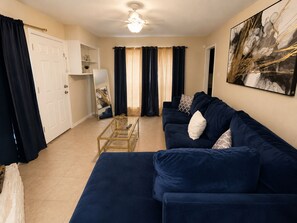 A living room with a dark blue sofa, a glass coffee table, a ceiling fan, and a large abstract painting on the wall. - Terrific 2-bedroom house minutes from Airport (Humble)