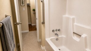 Un baño con una bañera blanca, un perchero con una toalla, una planta pequeña en un estante y una puerta que da a otra habitación.