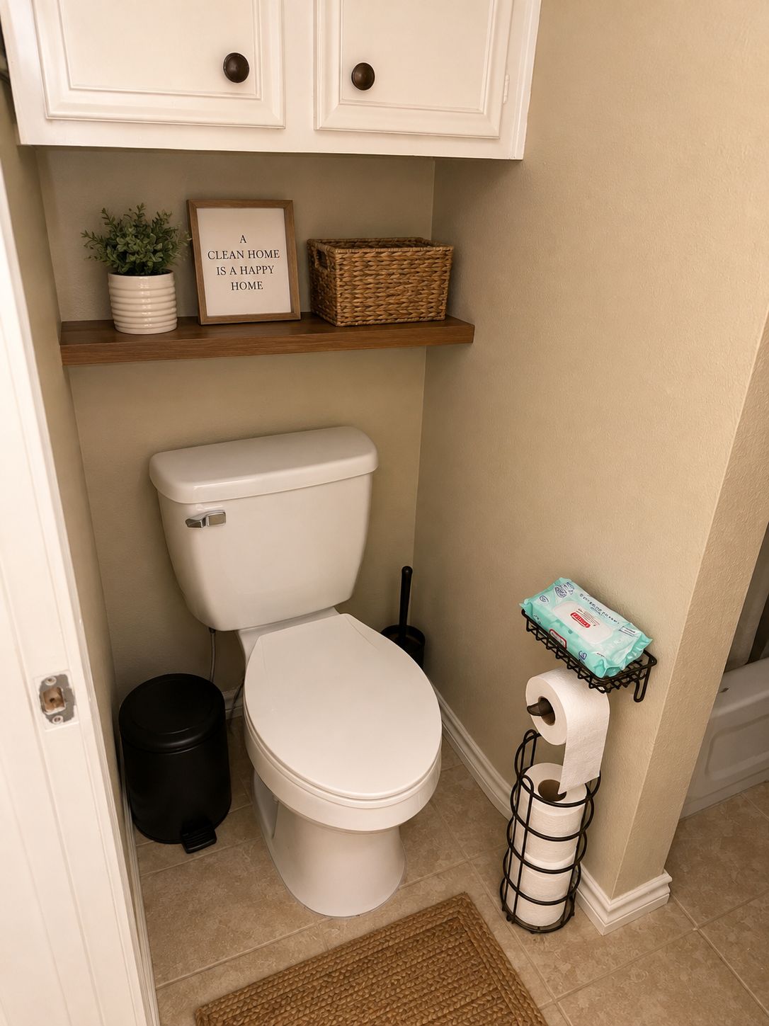 Ein sauberes Badezimmer mit Toilette, Mülleimer, Toilettenpapierhalter, einer kleinen Ablage mit Pflanze und gerahmtem Spruch sowie einer Tür.
