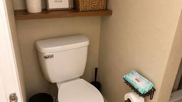 Ein sauberes Badezimmer mit Toilette, Mülleimer, Toilettenpapierhalter, einer kleinen Ablage mit Pflanze und gerahmtem Spruch sowie einer Tür.