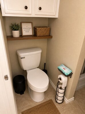 A clean bathroom with a toilet, a trash can, a toilet paper holder, a small shelf with a plant and a framed quote, and a door.