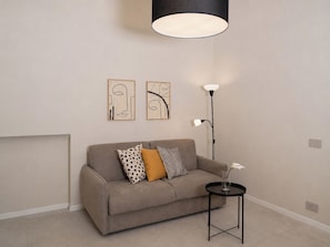 Ein neutral eingerichtetes Wohnzimmer mit einer Couch, zwei abstrakten Wandbildern, einer Stehlampe und einem kleinen Tisch mit einer Vase.