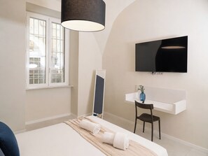 Quarto de hotel com cama, escrivaninha, cadeira, televisão e janela.