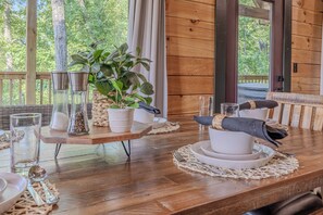 Una mesa de comedor de madera dispuesta con platos, cuencos y cubiertos blancos, junto con una planta y recipientes de vidrio sobre un aparador.