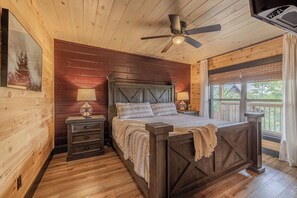 Un dormitorio de cabaña de madera con cama, mesitas de noche, ventilador de techo y ventana con vistas.