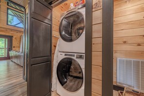 A laundry room with two stacked front-loading washing machines, wooden walls, and a large window. - Bear Bungalow | Mtn & Forest Views | Hot Tub (Sevierville)
