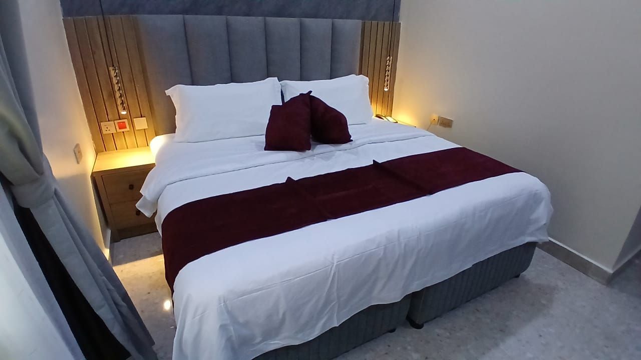 Una cama bien hecha con ropa blanca y una manta roja, una mesita de noche de madera con una lámpara y un cabecero tapizado en gris.