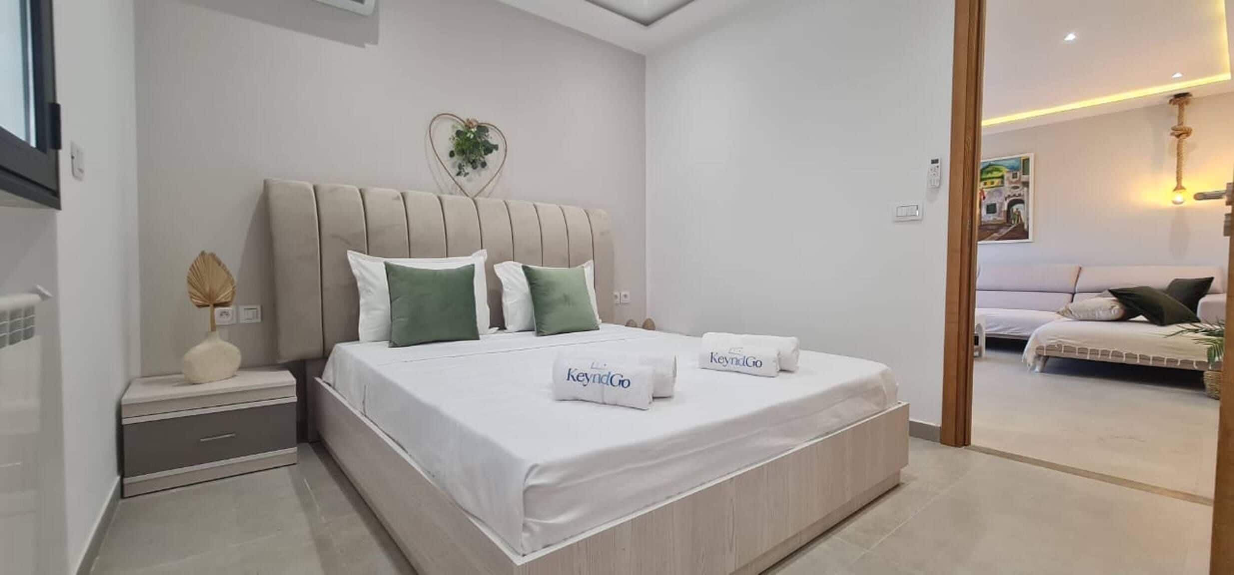Quarto moderno com cama grande, mesa de cabeceira, um detalhe decorativo em formato de coração e vista para outro cômodo.