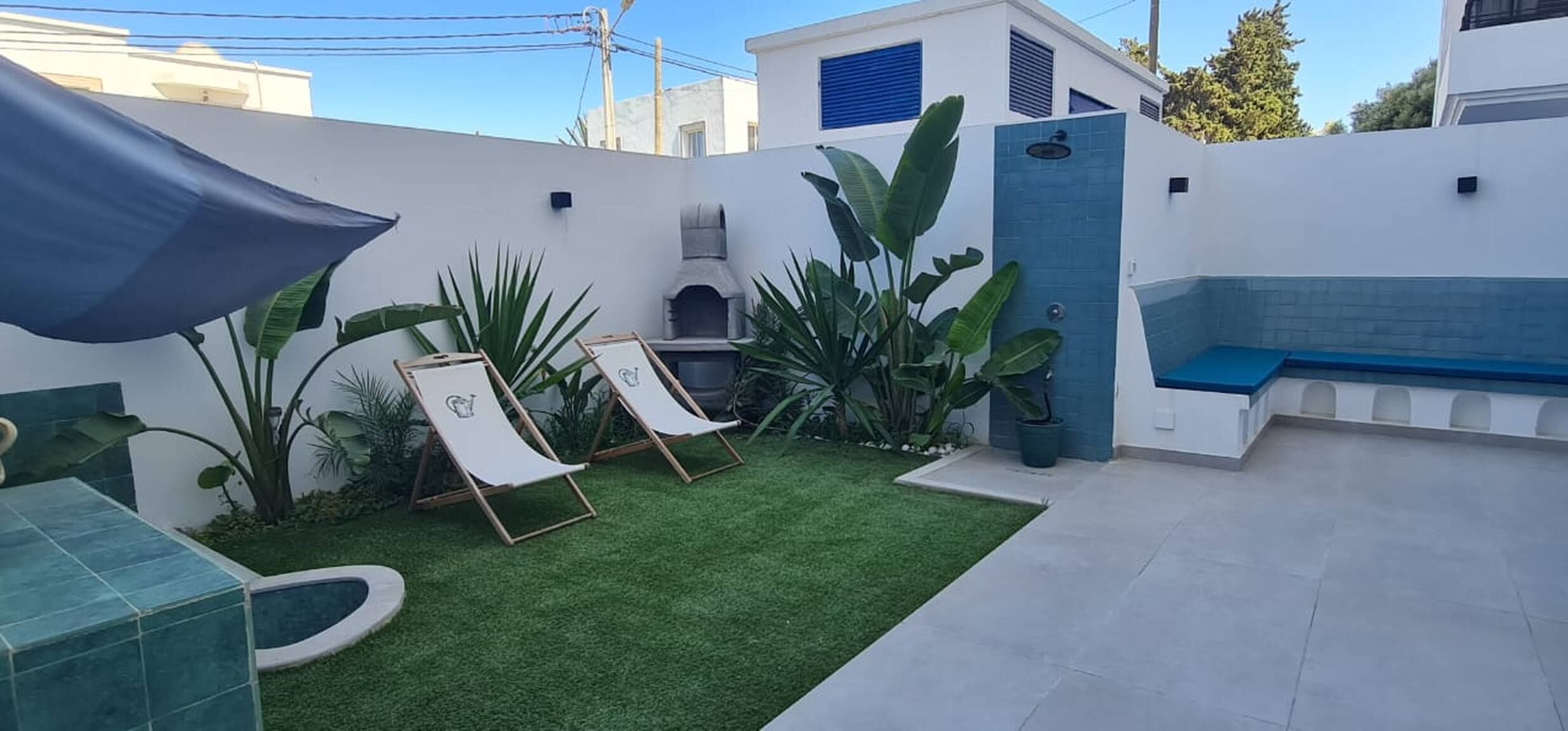 Uma área de pátio com grama sintética, duas poltronas de descanso, uma pequena piscina e um banco.