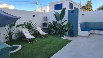 Uma área de pátio com grama sintética, duas poltronas de descanso, uma pequena piscina e um banco.