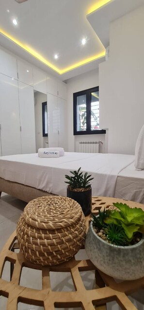 Quarto moderno com cama, mesa de cabeceira com planta e um banquinho de palha.