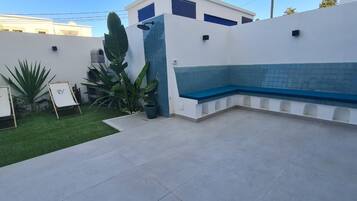 A patio area with a blue bench, a small garden with green plants, and a white wall with a window.