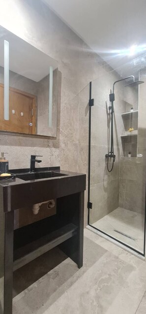 Casa de banho com toucador escuro, espelho, cabine de duche e chão em mosaico.