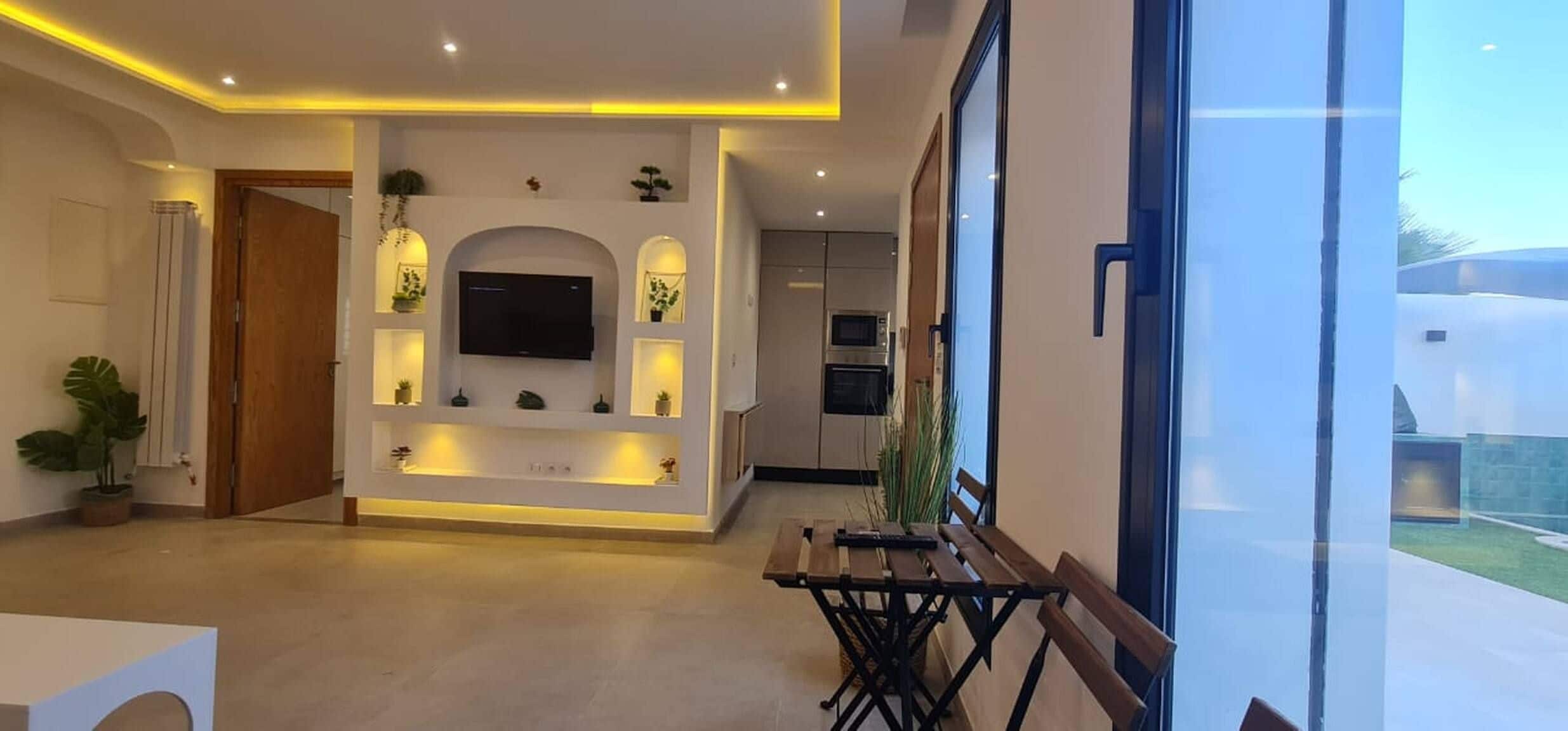 Un espace de vie moderne doté d’un téléviseur à écran plat fixé au mur, d’un éclairage encastré, d  ’une table à manger avec des bancs, et offrant une vue sur l’extérieur grâce à de grandes fenêtres.