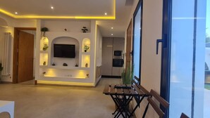 Un espacio de sala moderno con un televisor de pantalla plana montado en la pared, iluminación empotrada, una mesa de comedor con bancos y vistas al exterior a través de grandes ventanales.