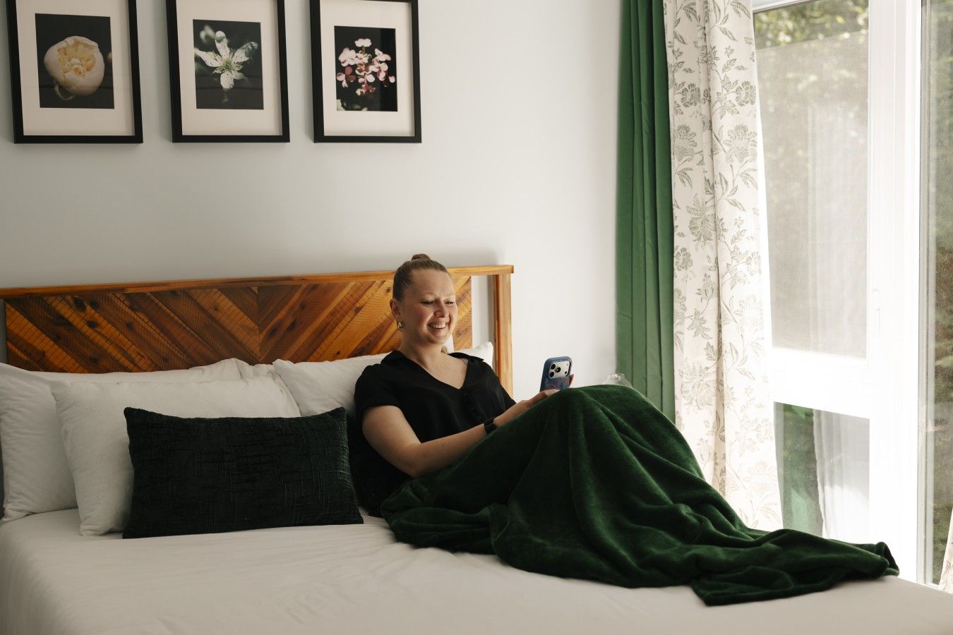 En person sidder på en seng med en smartphone, et træsenggærde og billeder i rammer på væggen.