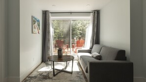 Un salon avec un canapé gris, une table basse ronde et une vue sur une terrasse extérieure avec des chaises rouges.
