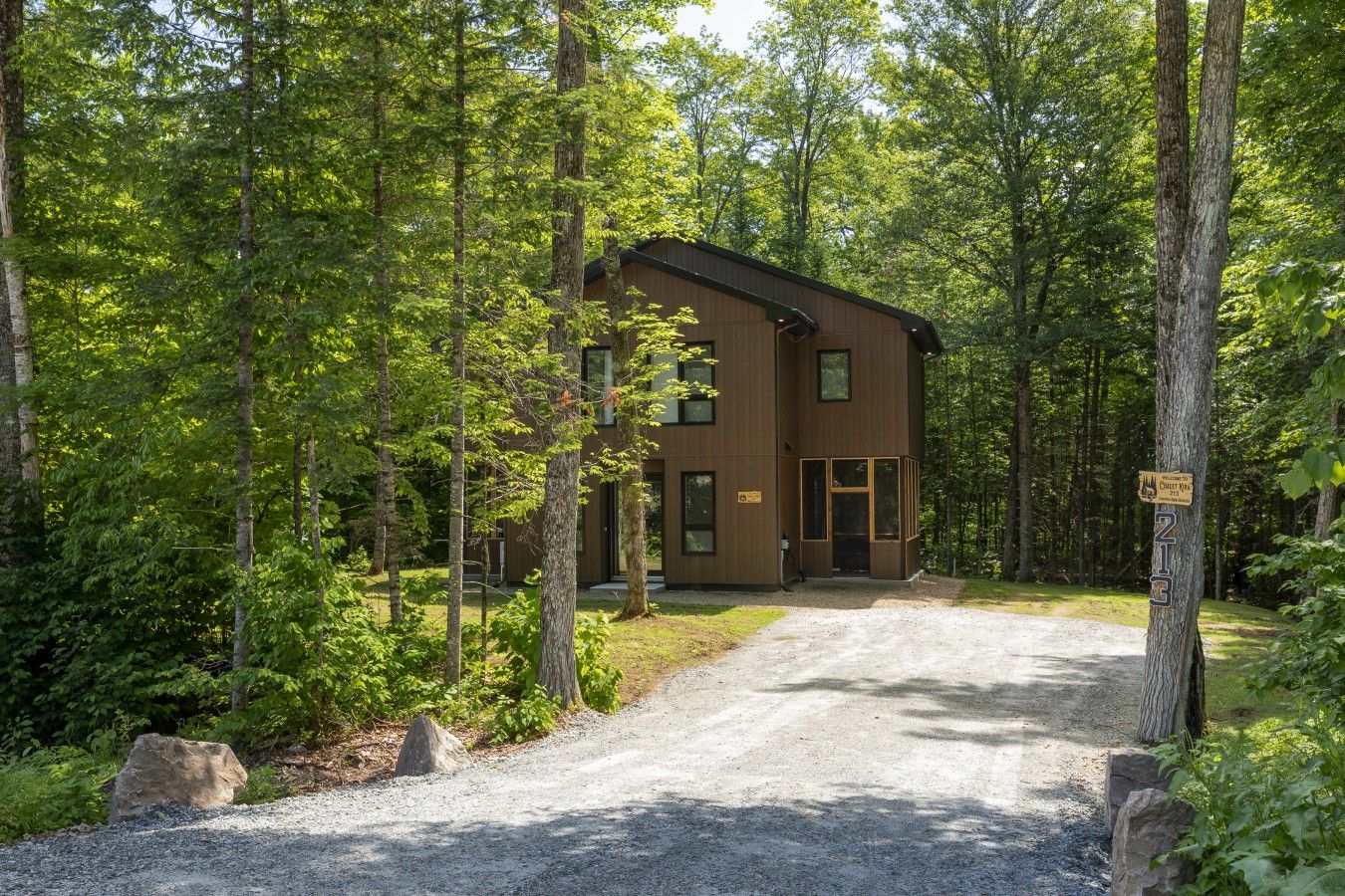 A two-story cabin with a gravel driveway leading to a wooden door, surrounded by dense forest.