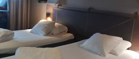 Habitación de hotel con dos camas, cada una con sábanas y almohadas blancas. Hay una lámpara roja en la pared, y el cuarto tiene un diseño moderno y sencillo.