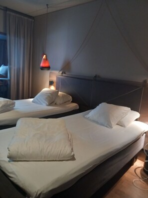 Ein Hotelzimmer mit zwei Betten, jeweils mit weißen Bettwäsche und Kissen, einem Nachttisch mit Lampe und einem Fenster mit Vorhängen.