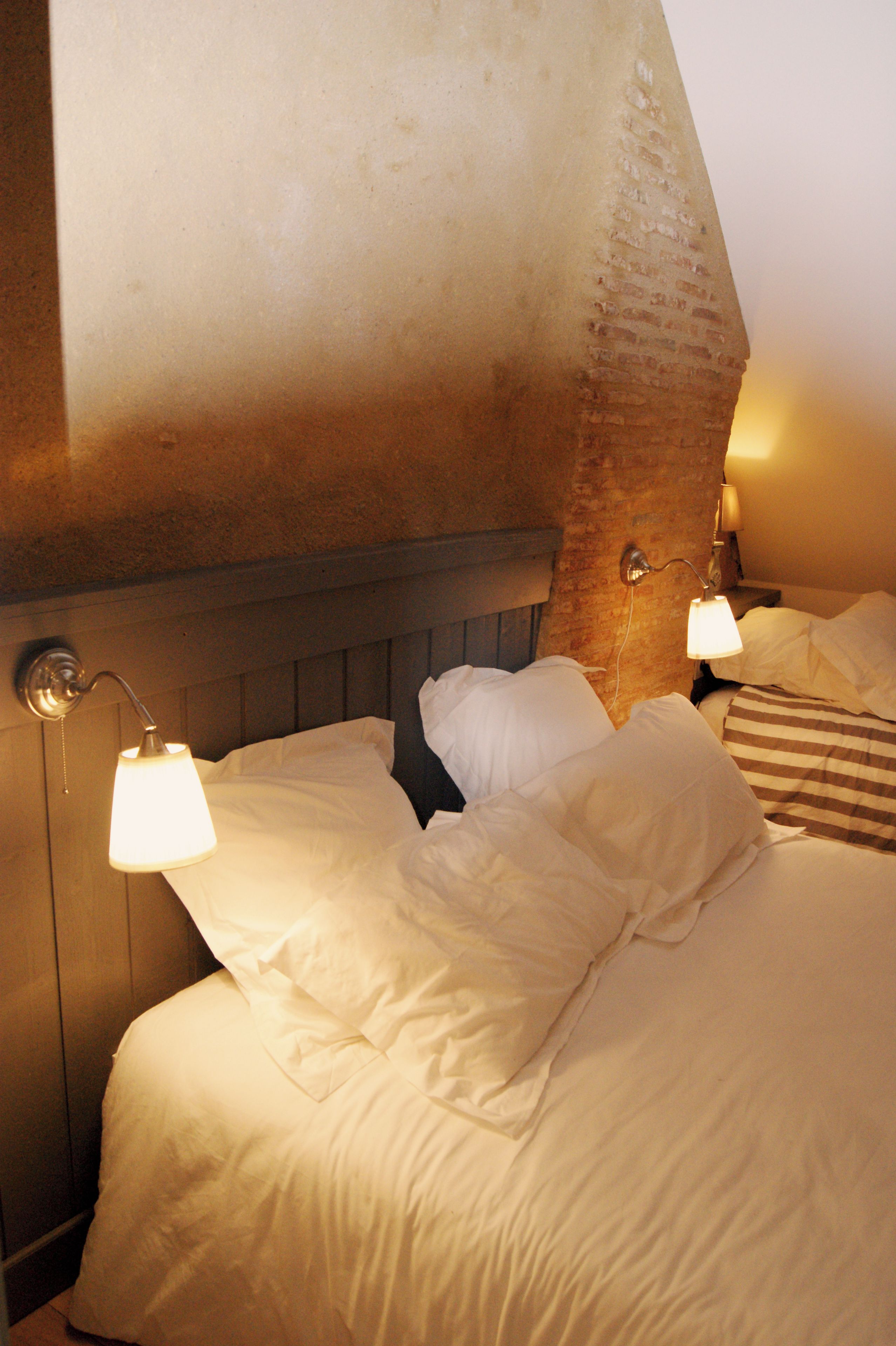 A hotel room with a bed, two bedside lamps, and a brick wall.