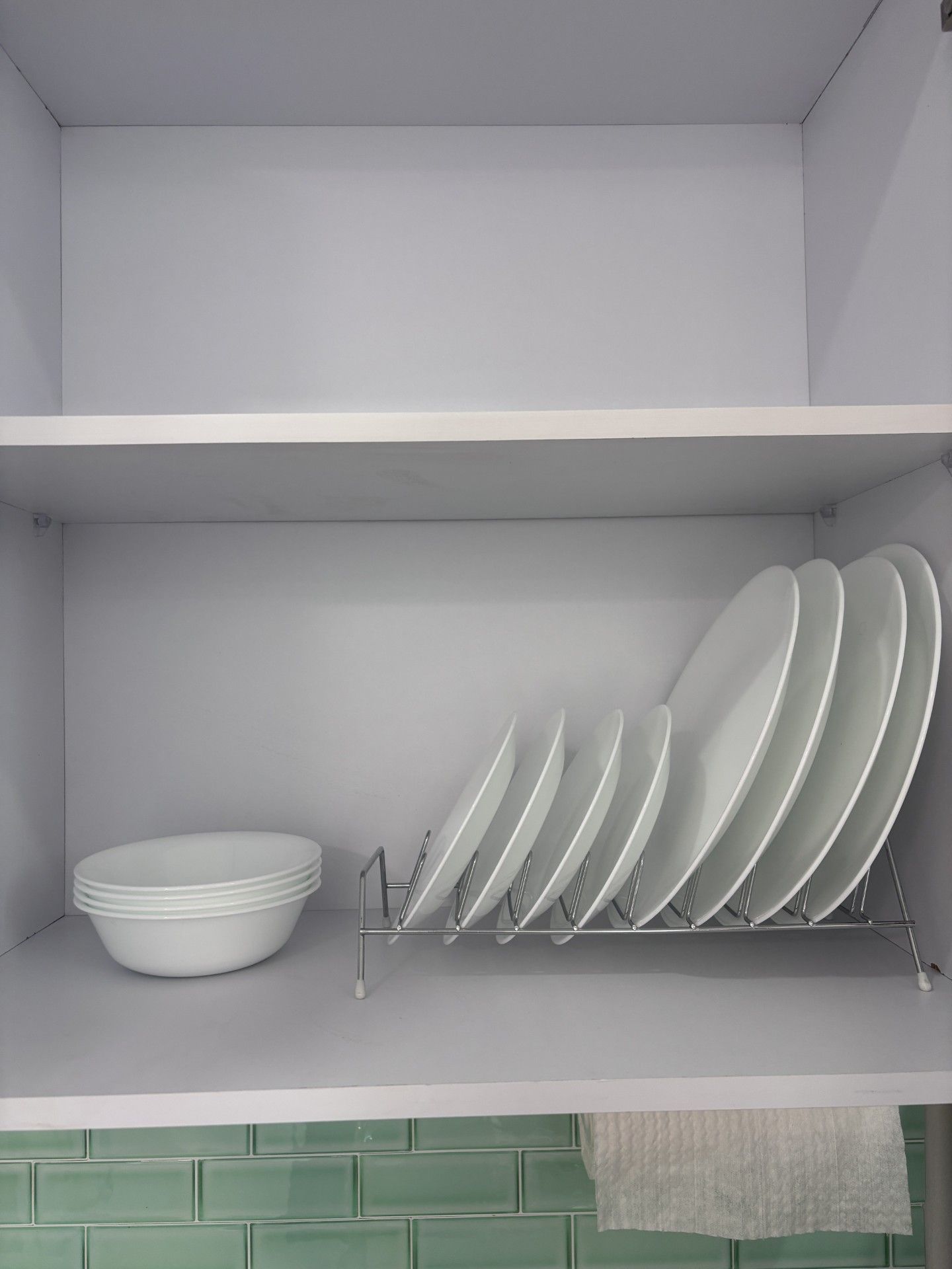 Una cocina con un estante blanco que contiene platos y tazones blancos, y un salpicadero de azulejos verdes.