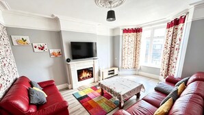 A living room with a fireplace, red sofas, a flat-screen TV, and a colorful rug. - South East London 5-bed House, Sleeps 7 Foresthill (London)