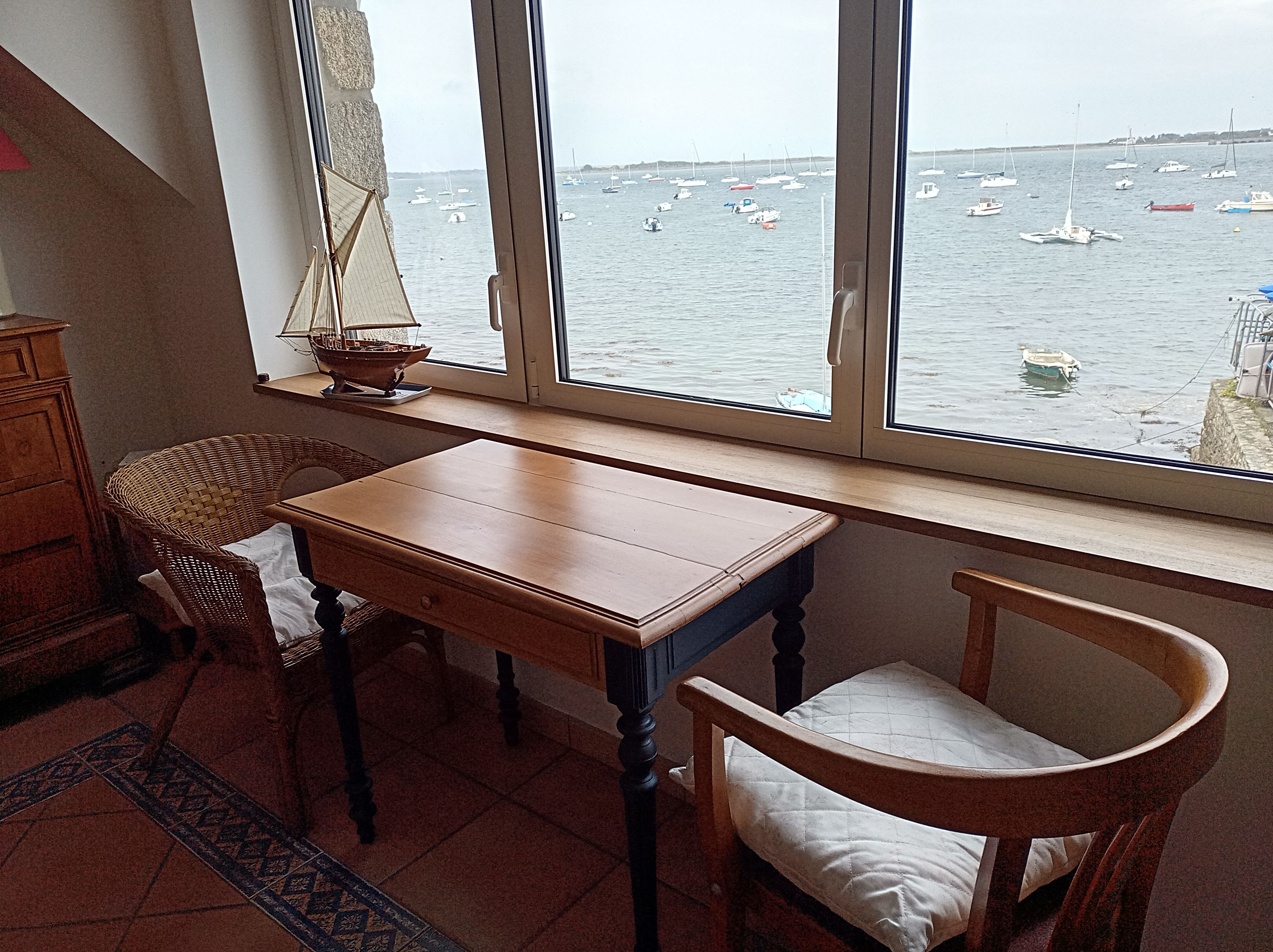 A room with a view of the sea, a wooden table, two chairs, and a small ship model.