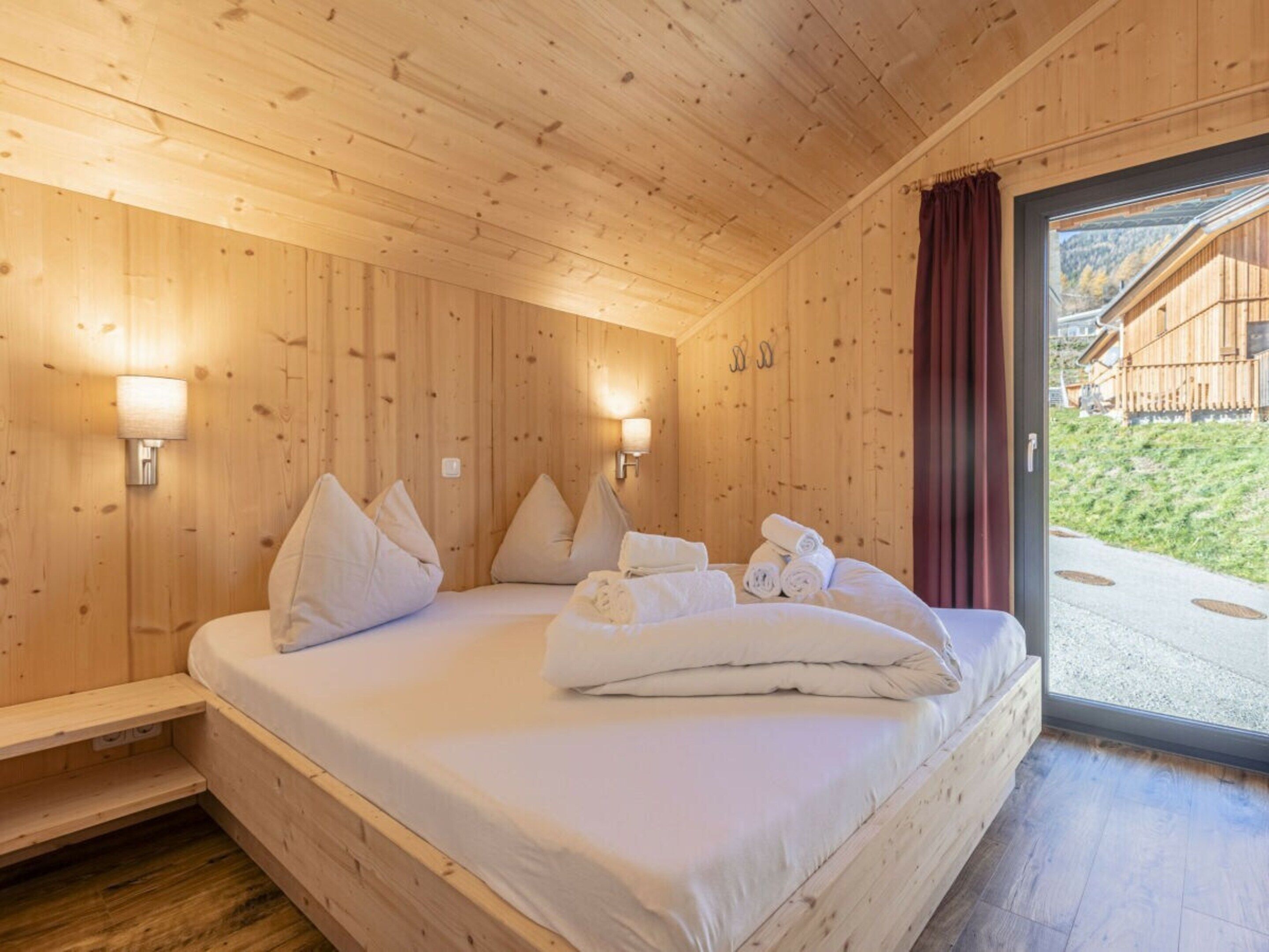 Ein Zimmer aus Holz mit einem Bett, Kissen und Handtüchern. An den Wänden sind Lampen angebracht und ein Fenster bietet Blick ins Freie.