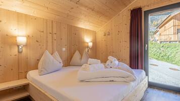 Ein Zimmer aus Holz mit einem Bett, Kissen und Handtüchern. An den Wänden sind Lampen angebracht und ein Fenster bietet Blick ins Freie.