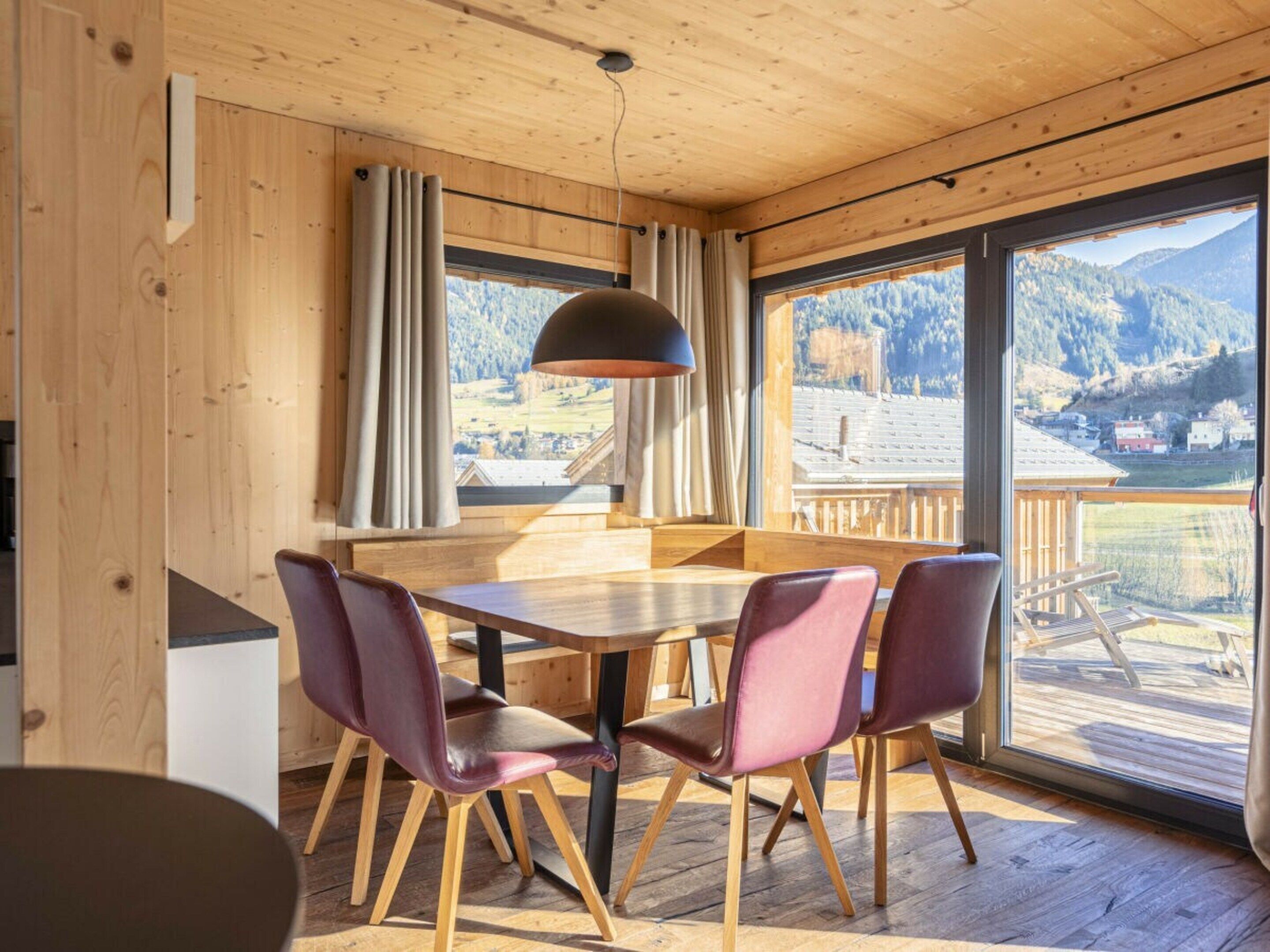 Un intérieur en bois avec une table et des chaises, de grandes fenêtres offrant une vue sur les montagnes et un luminaire suspendu.