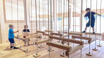 A wooden play structure with swings and climbing elements in a room with wooden flooring and large windows.