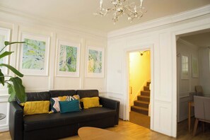 A living room with a black leather sofa, yellow and teal cushions, and framed abstract art on the wall. - Cozy 3-Bedroom Flat - Fitzrovia & Soho (London)