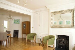A room with a fireplace, two green armchairs, a wooden cabinet, and a dining table. - Cozy 3-Bedroom Flat - Fitzrovia & Soho (London)