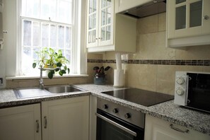 A kitchen with white cabinets, a granite countertop, a stove, an oven, a microwave, and a window with a plant. - Cozy 3-Bedroom Flat - Fitzrovia & Soho (London)