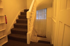 A staircase with carpeted steps and white balusters, leading to a landing with a door and a window with sheer curtains. - Cozy 3-Bedroom Flat - Fitzrovia & Soho (London)
