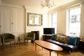 A living room with a sofa, two armchairs, a coffee table, a fireplace, a television, and large windows. - Cozy 3-Bedroom Flat - Fitzrovia & Soho (London)